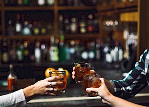 Three hands holding drinks across a bar counter, with shelves stocked with bottles in the background, inside a dimly lit bar.
