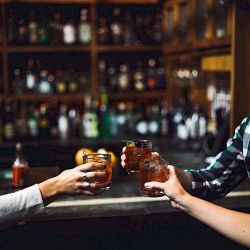 Three hands holding drinks across a bar counter, with shelves stocked with bottles in the background, inside a dimly lit bar.