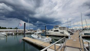 A marina filled with various boats and yachts under a dramatic cloudy sky; an American flag is visible on a pole to the left.