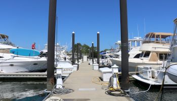 A marina with several boats docked, mooring lines, utility posts, and a clear, sunny sky.