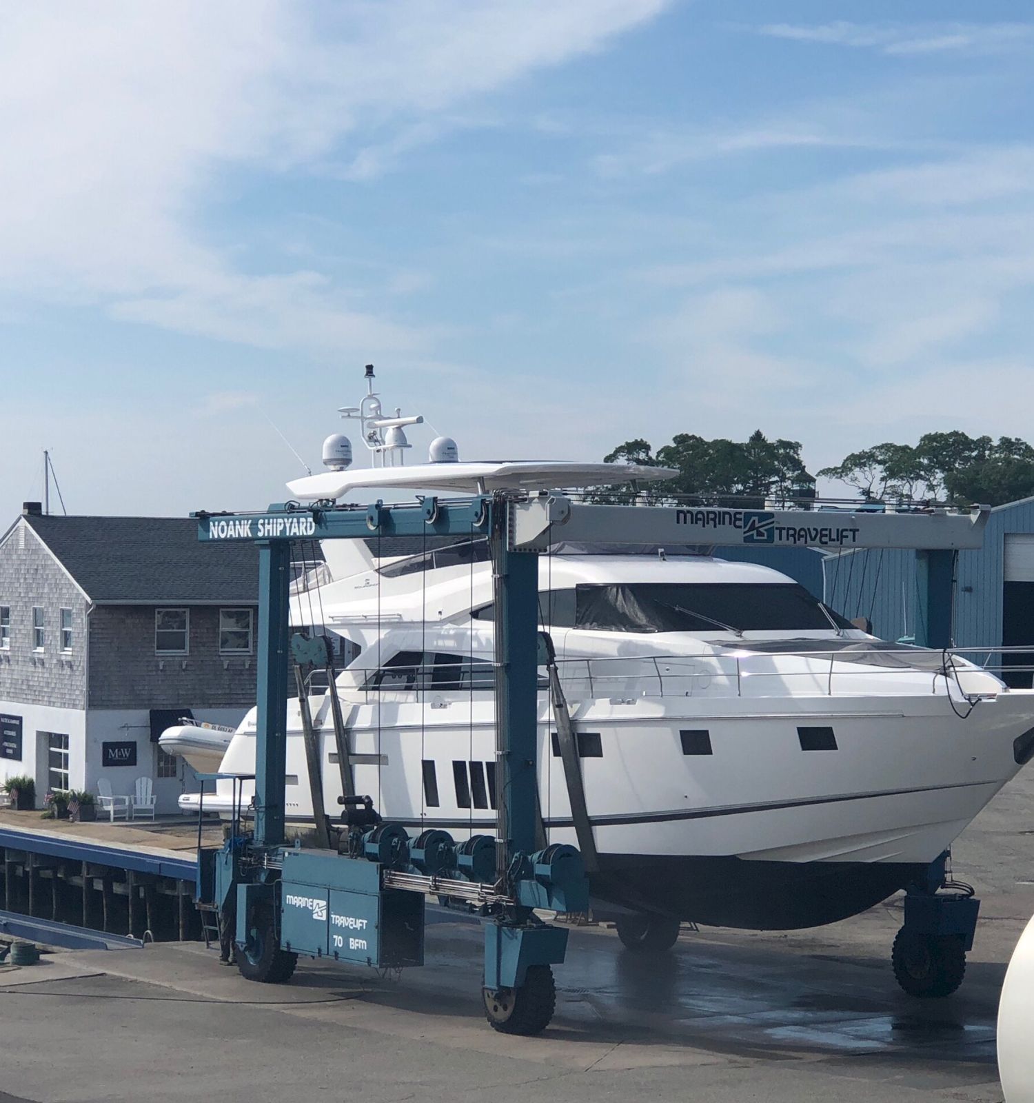 A boatyard with a large yacht being transported by a boat lift, surrounded by other boats, a building, and a scenic waterfront backdrop.