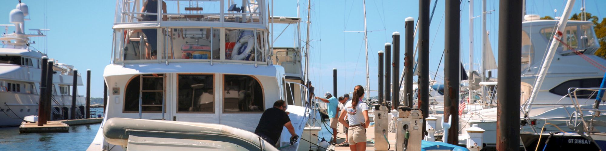 Several boats are docked at a marina. People are seen on the dock near one of the vessels, engaged in various activities. The sky is clear.