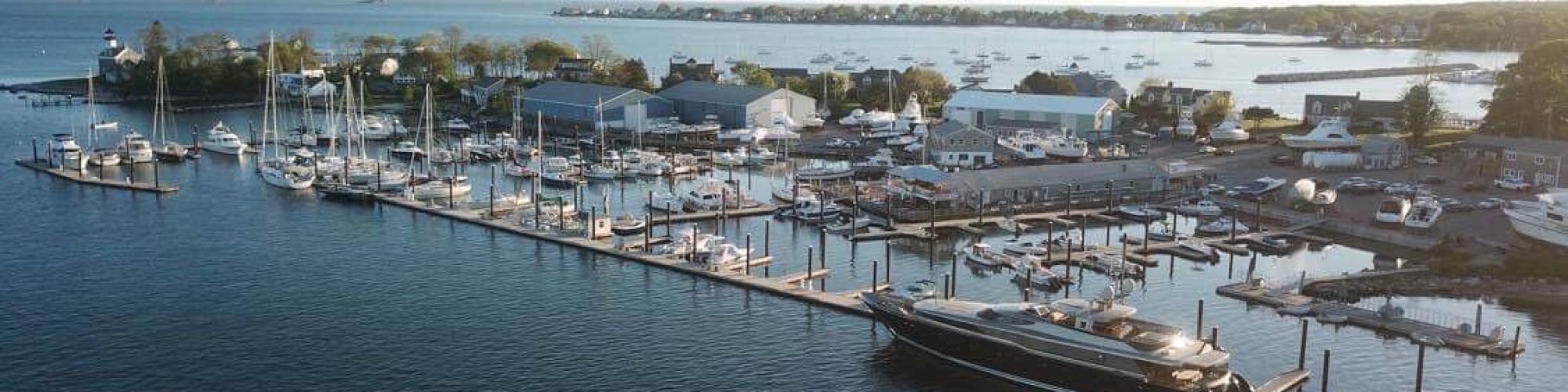 The image shows a marina with various boats docked at a long pier, buildings and trees in the background, and calm water in the foreground.