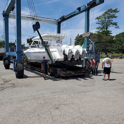 A boat is being lifted by a large crane-like vehicle in a yard with a few people standing nearby on a sunny day.