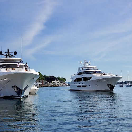 Two large yachts are docked in a calm harbor with clear blue skies and a few buildings in the background.