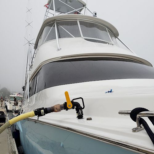 A close-up of a docked boat with a yellow fuel hose connected to it on a foggy day. The boat has multiple decks and antennae.