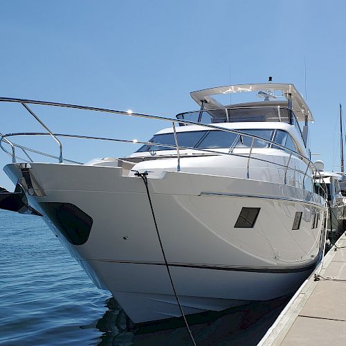 A large white yacht is docked at a marina under a clear blue sky, secured with ropes and surrounded by calm waters.