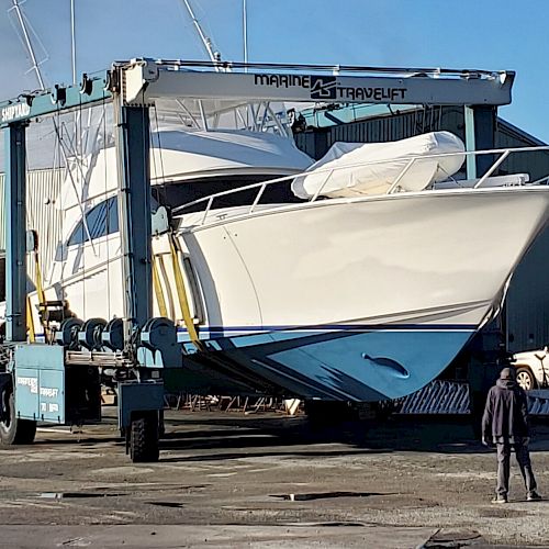 A large yacht is being transported by a Marine Travelift on land with one person standing nearby, viewing the process.