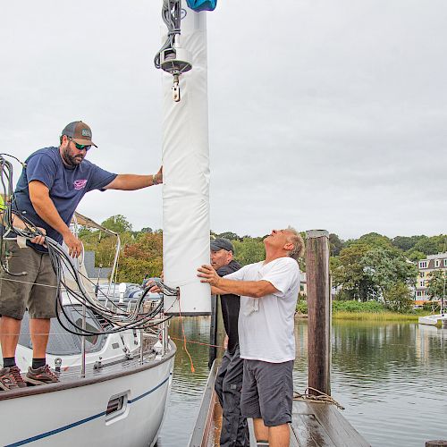 Several people are working on or around a sailboat at a dock, possibly preparing the mast or sail, with houses and water in the background.