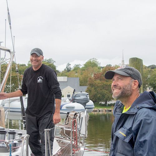 Two men on a boat at a marina, smiling and dressed in casual outdoor attire, with buildings and trees in the background.