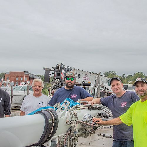 Five men stand together outdoors near a large piece of equipment, with boats and buildings in the background.