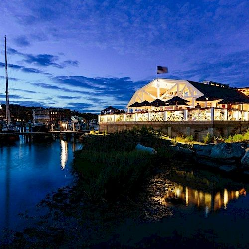 A serene waterfront scene at dusk, with a well-lit building, a docked boat, and a calm body of water reflecting the evening sky, ending the sentence.