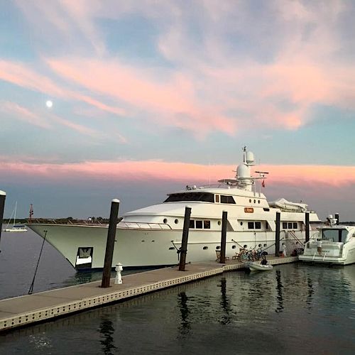 A luxury yacht docked in a marina at sunset with a pink and blue sky in the background and a few smaller boats beside it.