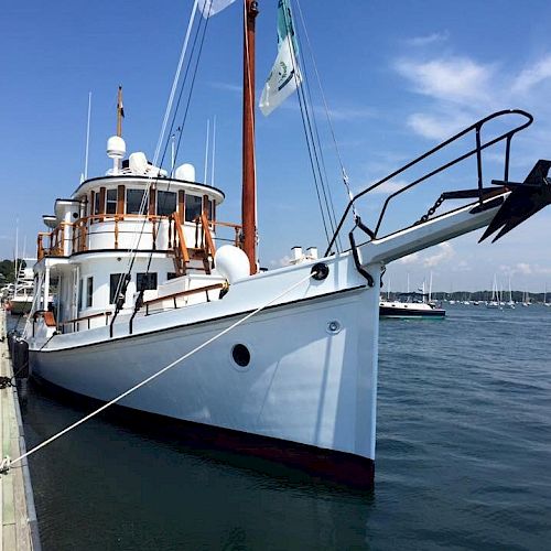 A white boat is docked at a marina under a clear blue sky, with several flags fluttering near its mast.