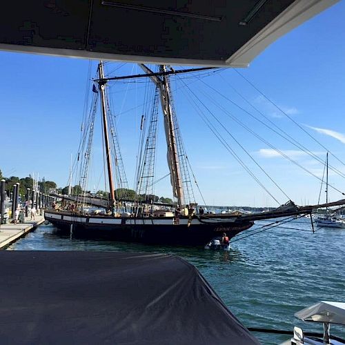 A tall ship is docked at a marina, with people on the pier and sailboats in the background, under a clear blue sky.