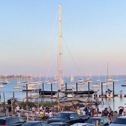 A busy harbor scene with many boats docked at the marina. People are gathered near the water, and cars are parked in the foreground.