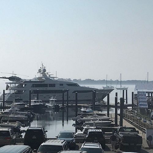 A marina with parked yachts and boats, cars along the sides, a large yacht with a helicopter on it, and a misty background.