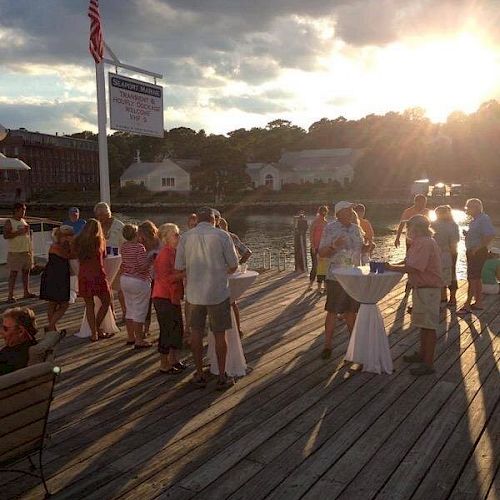 A group of people are gathered on a wooden dock near the water, socializing under a setting sun with an American flag raised, silhouetted by the sunlight.