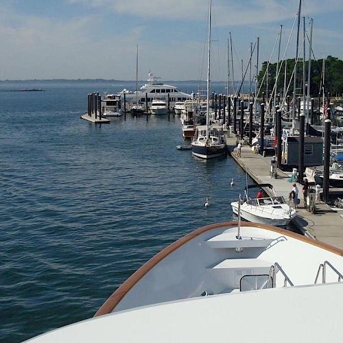 A marina with several boats docked along a pier extending into calm waters, with the bow of a large vessel in the foreground under a partly cloudy sky.