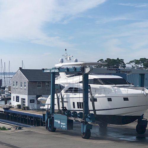 A large white yacht is being transported out of the water by a boat lift at a marina. There are docks, a building, and sailboats in the background.