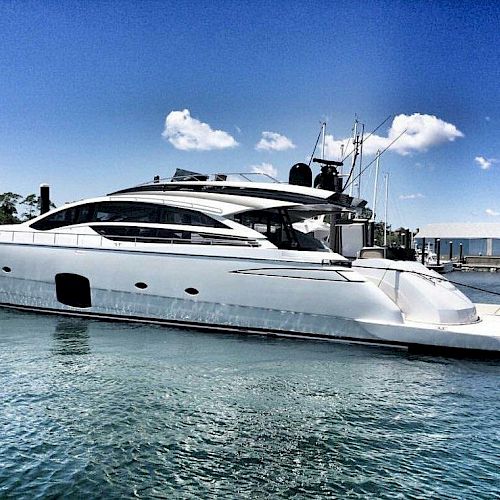 A sleek, modern white yacht is docked at a marina under a clear blue sky with some scattered clouds.