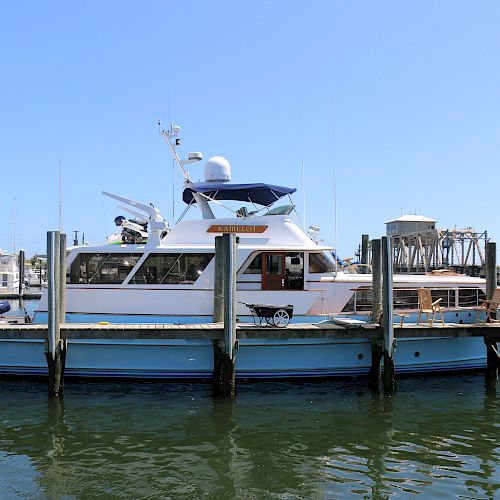 A white boat is docked at a pier on a sunny day, with calm water and a clear blue sky in the background.