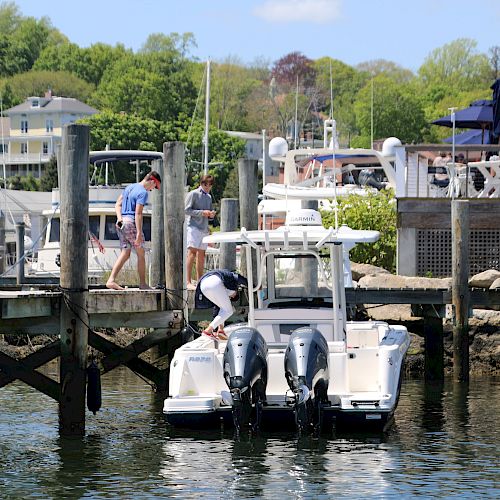 People are on a dock next to a white boat with dual outboard motors. Other boats and trees are visible in the background.