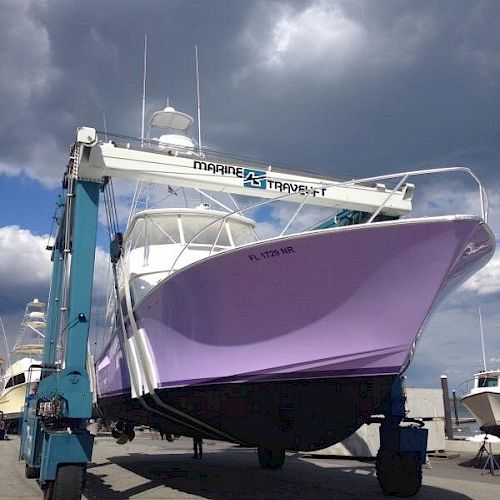 A purple boat is being lifted by a large crane-like machine at a marina, with a cloudy sky in the background.