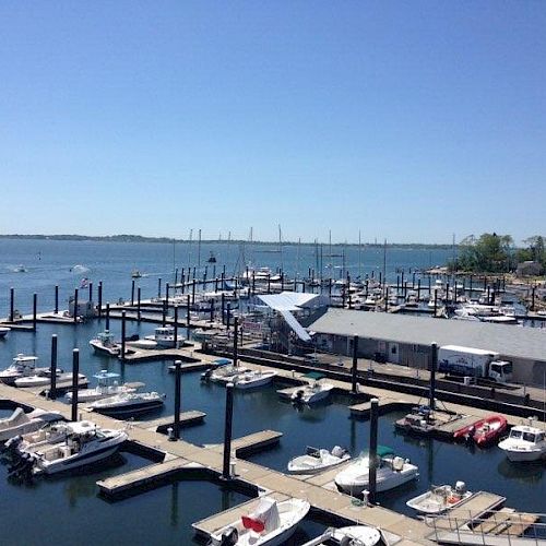 A marina with numerous boats docked, situated by a calm and expansive body of water under a clear blue sky.