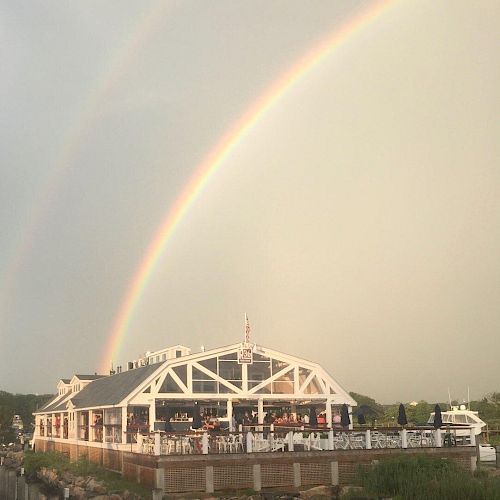 The image shows a waterfront building under a double rainbow with a cloudy sky in the background. The building appears to be a restaurant or café.