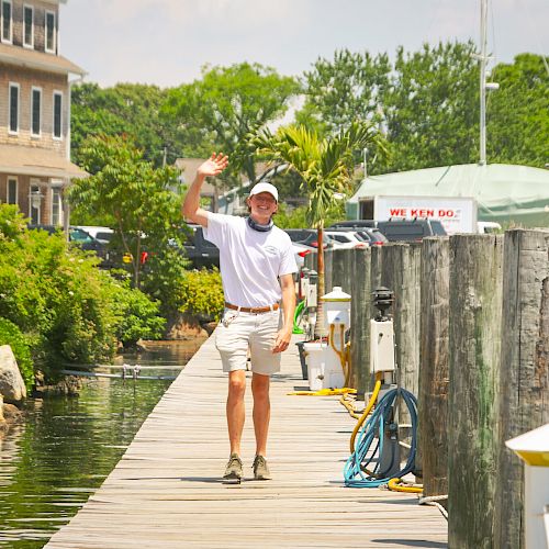 A person in a white outfit waves while walking on a sunny dock, with boats and a building in the background.