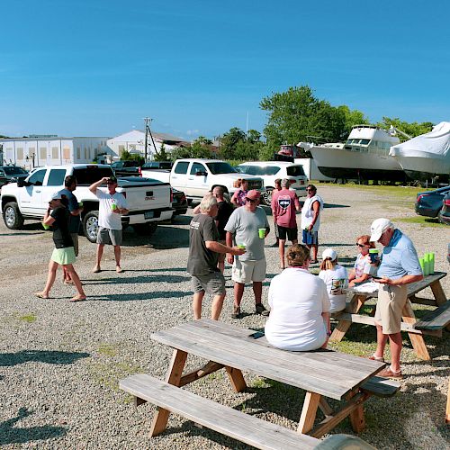 A group of people is gathered outdoors near a few parked vehicles and boats. Some are standing while others sit at picnic tables.