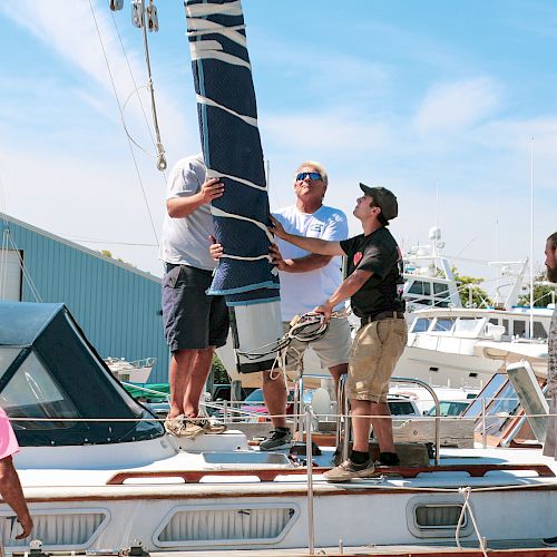 A group of people are working together to raise a sail on a docked sailboat at a marina on a clear day.