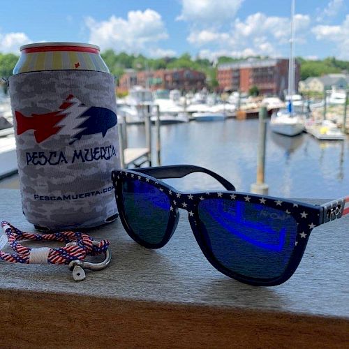 A can in a koozie, sunglasses, and a braided bracelet are on a ledge overlooking a marina on a sunny day.