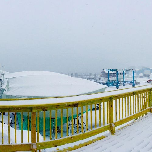 A snowy scene with boats covered in snow, viewed from a yellow wooden deck with a railing. The background dissipates into a foggy distance.