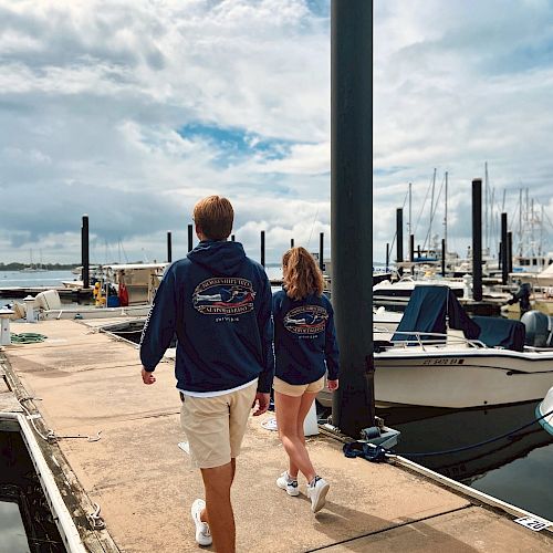 Two people in matching hoodies walk on a dock lined with boats under a cloudy sky, possibly near a marina.