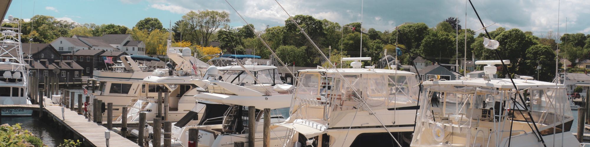 A marina with several docked boats, bright sky with scattered clouds, and green trees in the background. An American flag is visible in the scene.