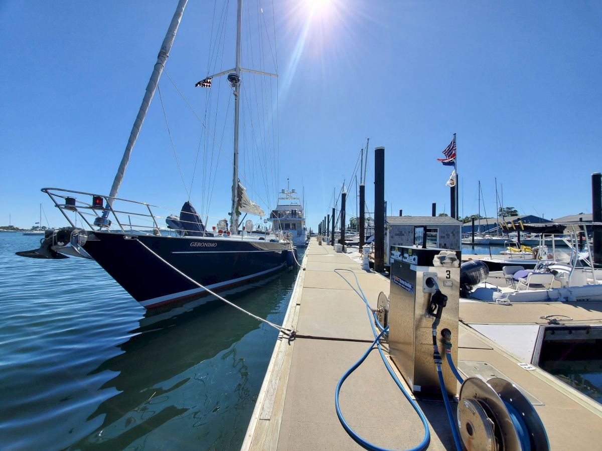 A sunny day at a marina with several sailboats and yachts docked along the pier, with flags waving and equipment on the dock.