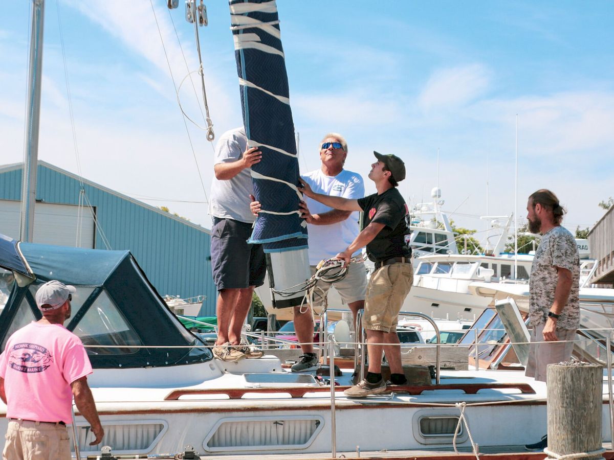 A group of people is on a docked boat, adjusting the sail and preparing the boat. The setting appears to be a marina with other boats in the background.