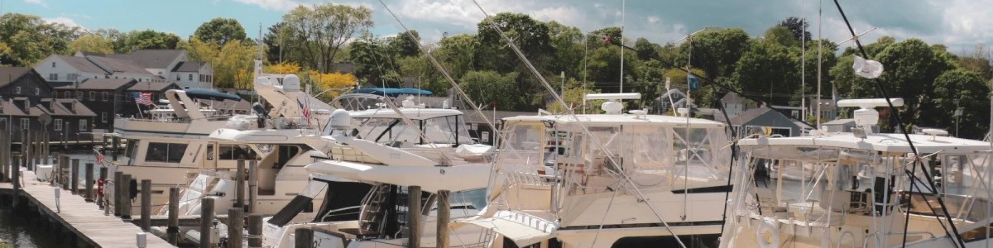 Several yachts are docked at a marina on a sunny day, with an American flag visible. There are green trees and houses in the background.