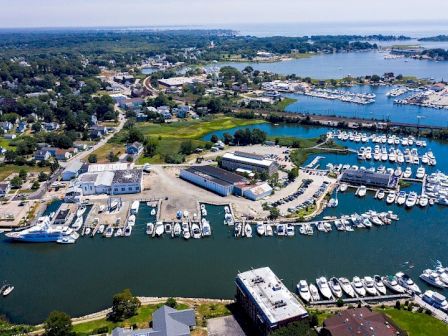 An aerial view of a coastal town with numerous boats docked in a marina, buildings, and greenery visible, while water surrounds the area.