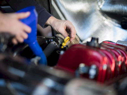 Two hands are working on a car engine; one pours fluid via a blue funnel while the other handles a component near a red engine part.