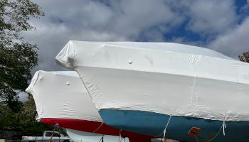 Two boats are covered with protective tarps, resting on stands against a partly cloudy sky.