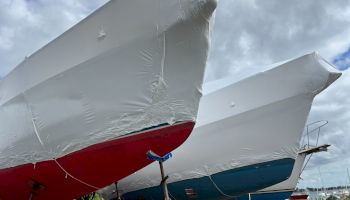 The image shows two boats in dry dock, wrapped in protective covers, with one having a red hull and the other a blue hull.