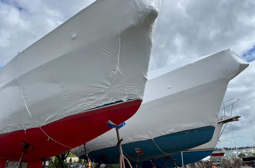 The image shows two boats in dry dock, wrapped in protective covers, with one having a red hull and the other a blue hull.